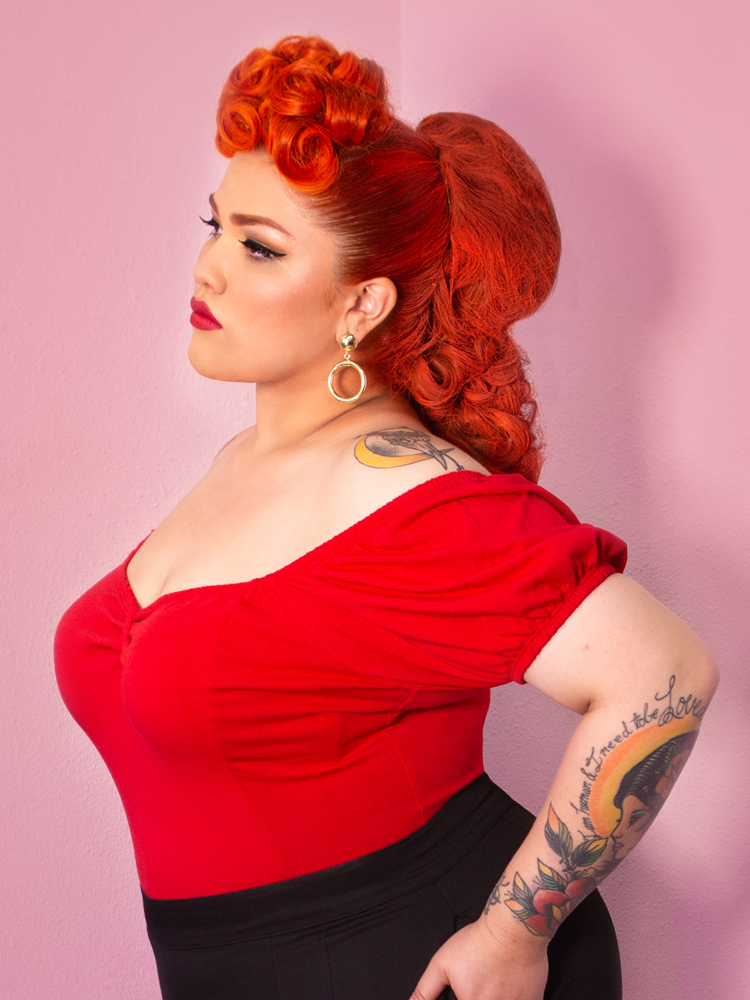 A side shot of Evelyn looking off camera with her hands on her hips modeling the Powder Puff top in red by Vixen Clothing.