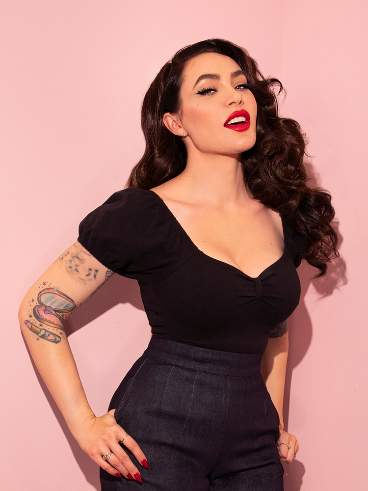 With her thumbs tucked into either of her pockets, Micheline Pitt smiles at the camera as she looks upward while wearing a classic outfit highlighted by the Powder Puff Top in Black.