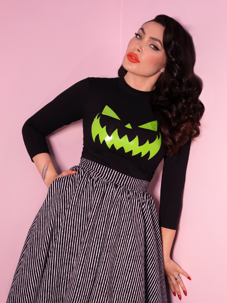 Standing and slightly leaning against a pink wall, Micheline Pitt models a retro style top featuring glow in the dark jack-o-lantern artwork that has been screen-printed on the front.