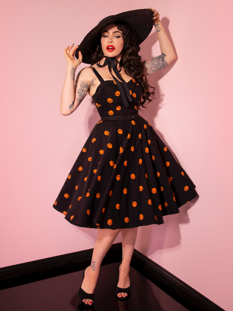 Placing her hands on the brim of her black sunhat, Micheline Pitt twirls in the Pumpkin King Maneater Swing Dress in Black.