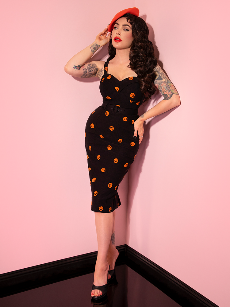 Posing with her hand on her hip and the other holding her orange beret, Micheline Pitt looks off-camera while wearing the Pumpkin King Maneater Wiggle Dress in Black.