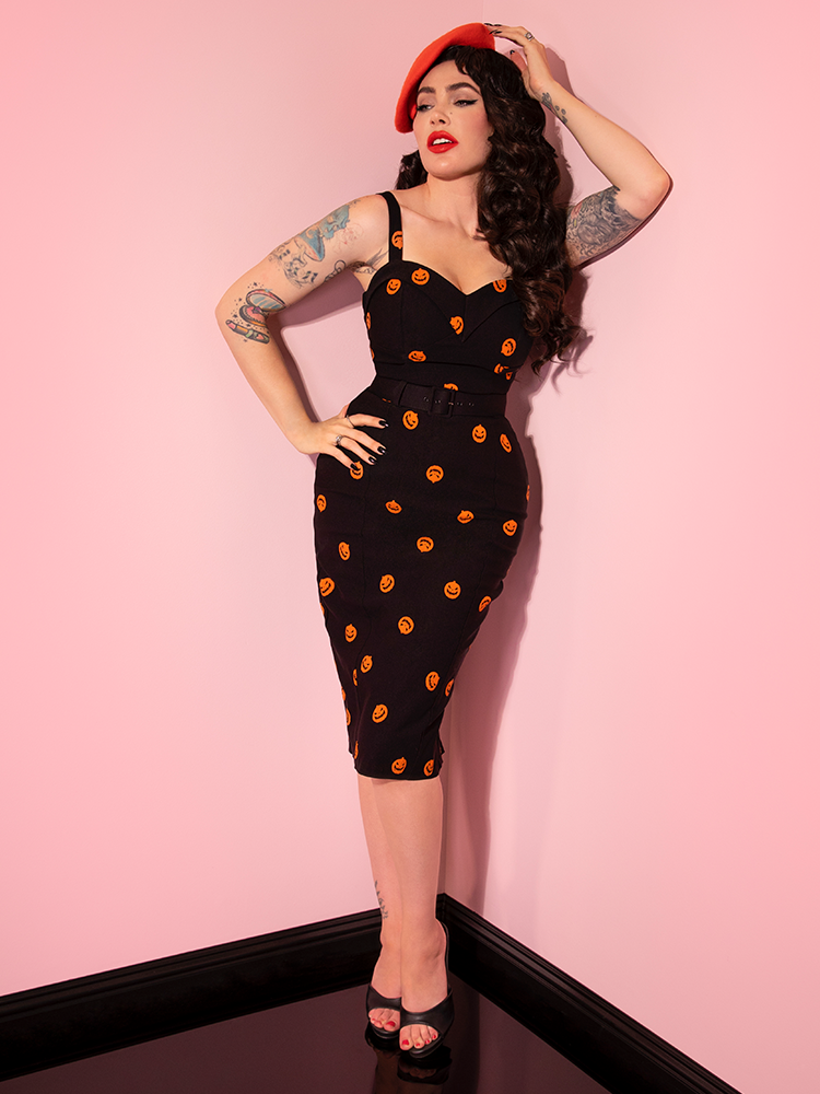 Micheline Pitt posing in her pink showroom while wearing the Pumpkin King Maneater Wiggle Dress in Black.