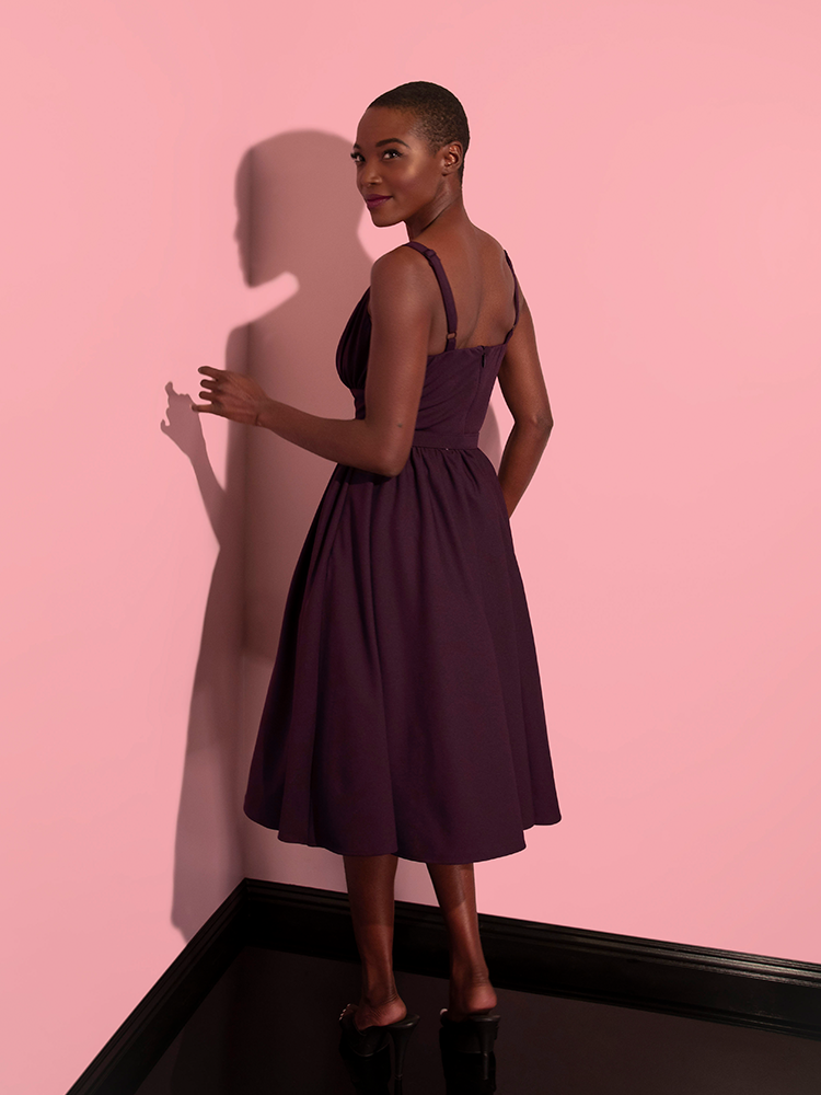 Model turned away from the camera but looking back over her shoulder while wearing the Ingenue Swing Dress in Eggplant Purple.