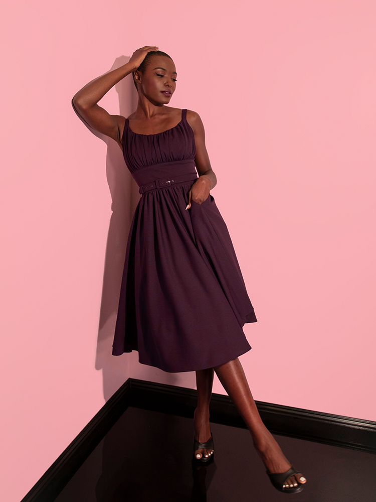 Model posing by leaning on the wall with one hand in her pocket and the other on top of her head while wearing the Ingenue Swing Dress in Eggplant Purple.