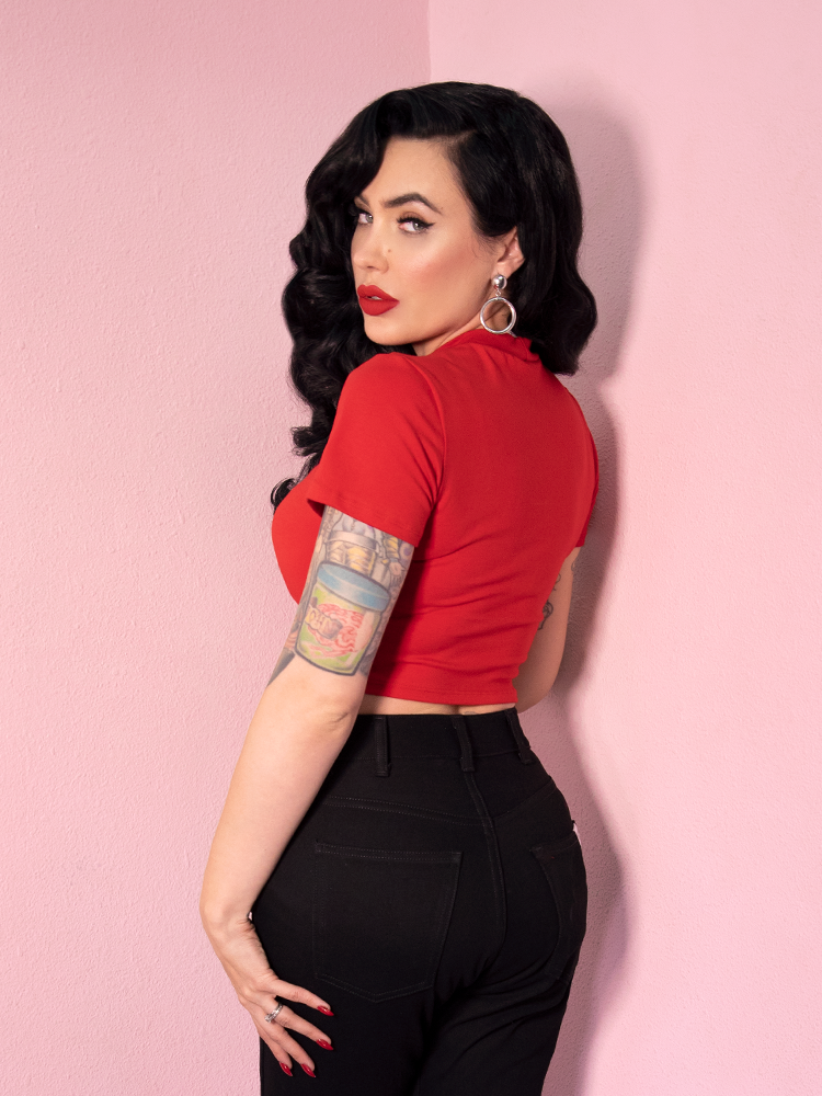 With her body turned away from the camera, Micheline Pitt looks back and over her shoulder at the camera to show off the Bad Girl Crop Top in Red from retro clothing company Vixen Clothing.