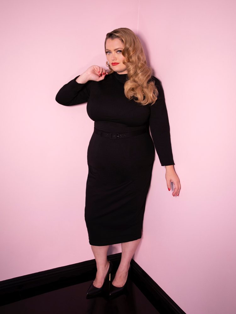 Blondie posing in the corner of a pink room with the Bad Girl Wiggle Dress in Black from retro dress retailer Vixen clothing.