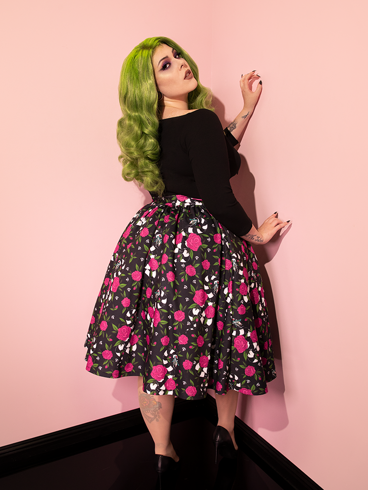 Green haired model turned away from the camera, but looking back over her shoulder at it, wears the BEETLEJUICE™ Sandworm & Roses Swing Skirt with a long-sleeve vintage style top.