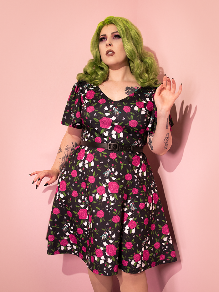 Green haired model posing delicately in the BEETLEJUICE™ Sandworm & Roses Swing Dress with matching black belt.