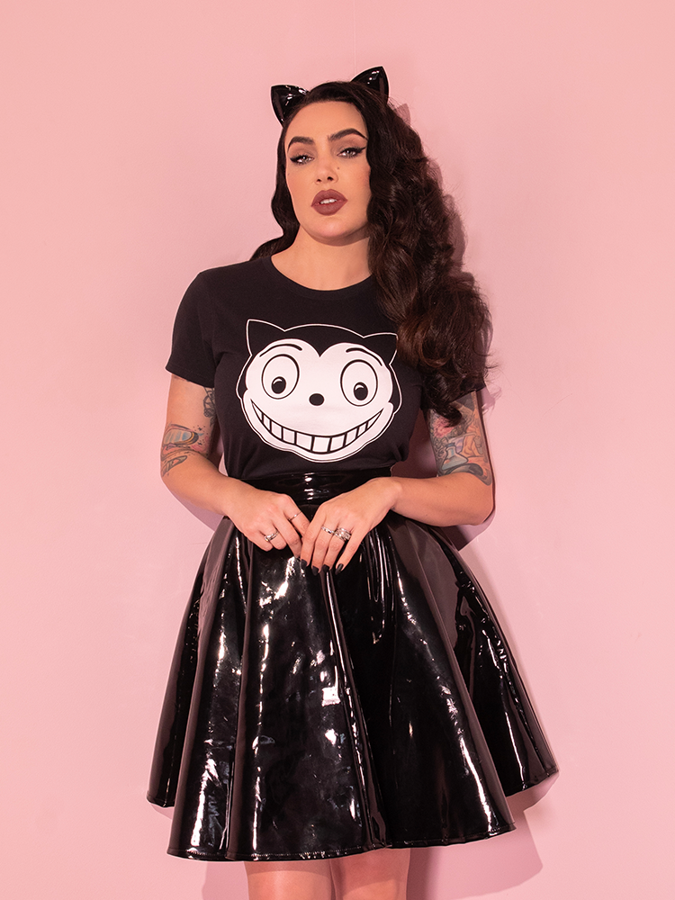 Micheline is standing in a pink room while wearing the Batman Returns Shreck's Cat women's tee and a vinyl skater skirt.