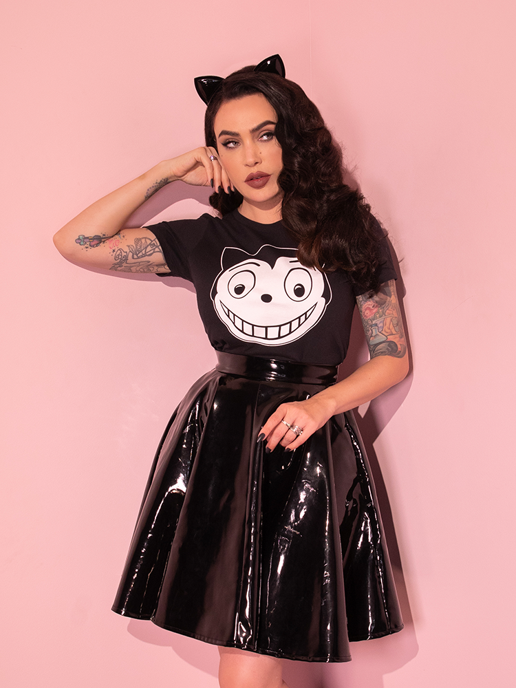 Micheline is standing in a pink room looking to the side while wearing the Batman Returns Shreck's Cat women's tee and a vinyl skater skirt.