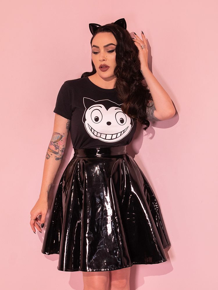Micheline is standing in a pink room with her hand in her hair while wearing the Batman Returns Shreck's Cat women's tee and a vinyl skater skirt.