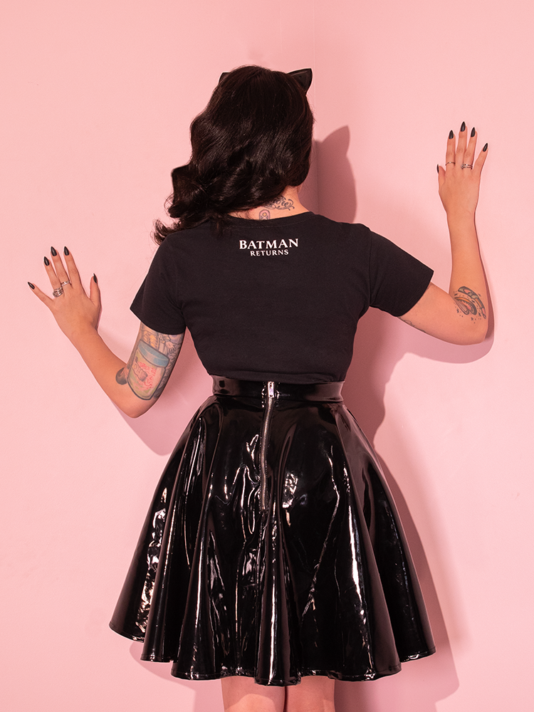 Micheline is facing the wall in a pink room while wearing the Batman Returns Shreck's Cat women's tee and a vinyl skater skirt.