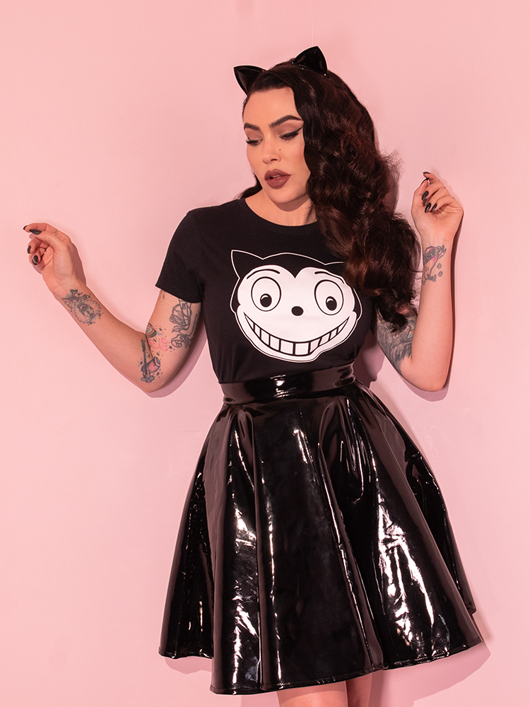 Micheline is standing in a pink room looking down while wearing the Batman Returns Shreck's Cat women's tee and a vinyl skater skirt.