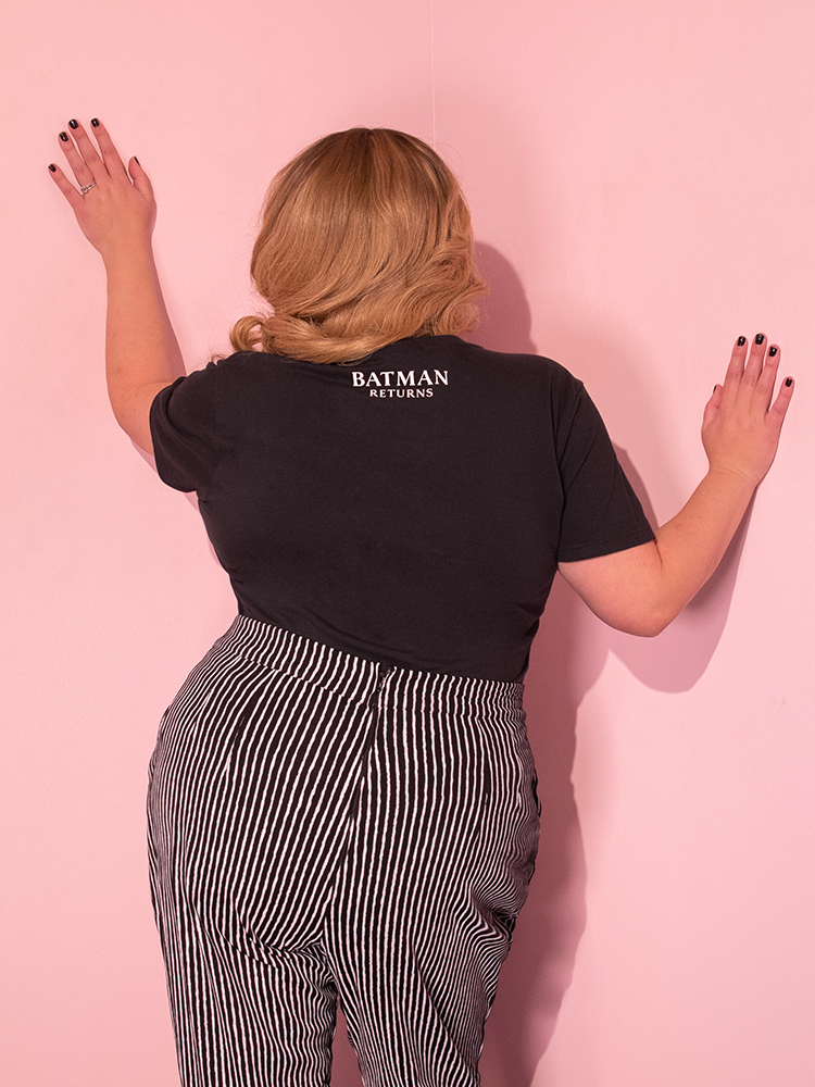 Blondie is facing the wall in a pink room while wearing the Batman Returns Shreck's Cat women's tee and high waisted pants.