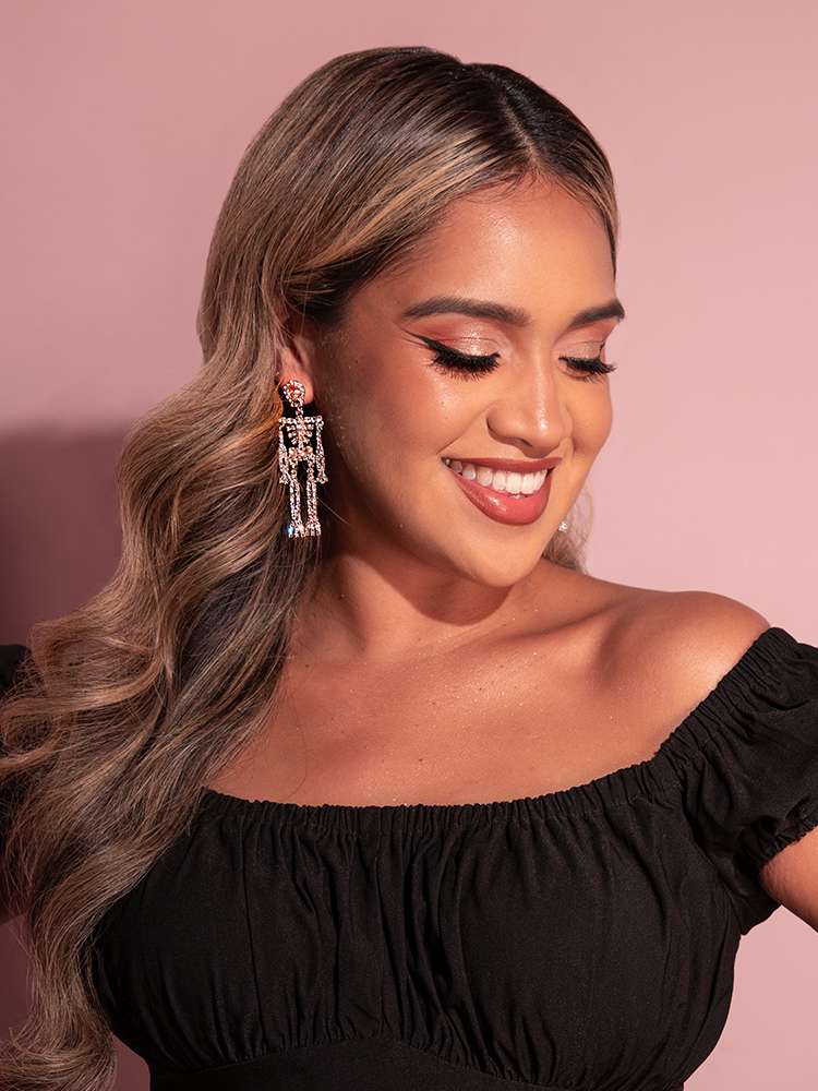 Model smiles while looking down and to the side while wearing the Rhinestone Skeleton Dangle Earrings in White from retro clothing brand Vixen Clothing.