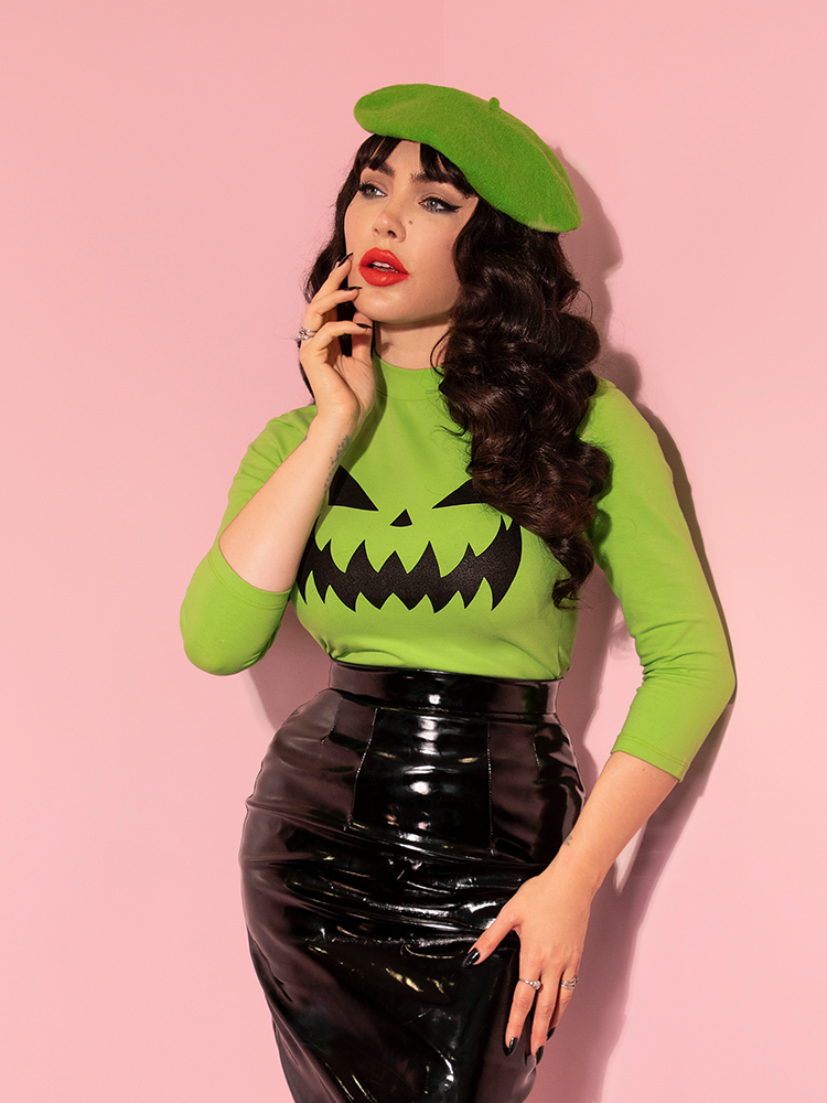 In a contemplative pose, Micheline Pitt models the Pumpkin King 3/4 Sleeve Top in Slime Green from Vixen Clothing.