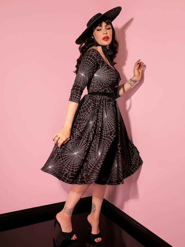 Micheline Pitt turned to the side while wearing the Wicked Swing Dress in Vintage Spiderweb Print from Vixen Clothing.