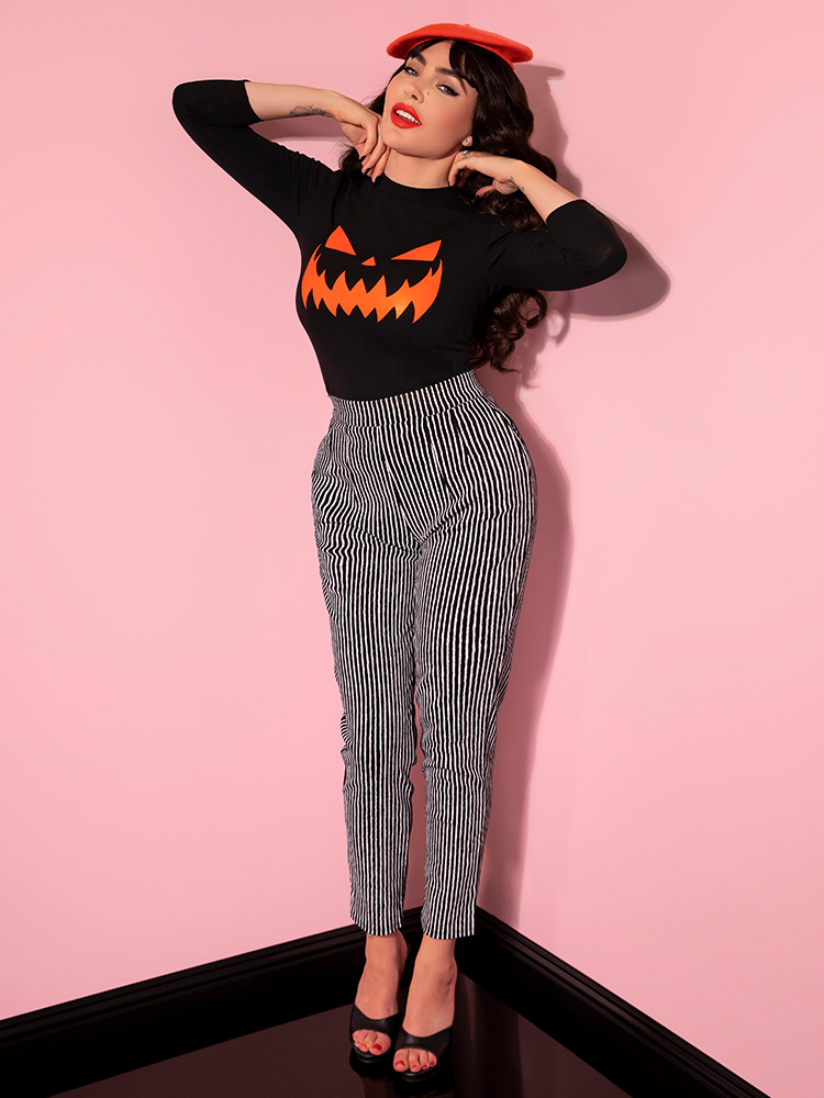 Smiling and looking into the camera with her hands on her shoulders, Micheline models the Miss Kitty Cigarette Pants in Black Stripes by Vixen Clothing.
