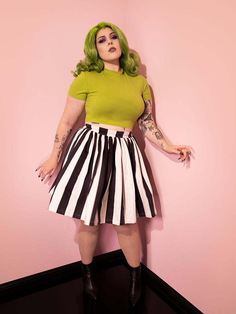 Model posing in the corner of a pink room wearing the Ghost Skater Skirt in Black & White Stripes and short sleeve green top.
