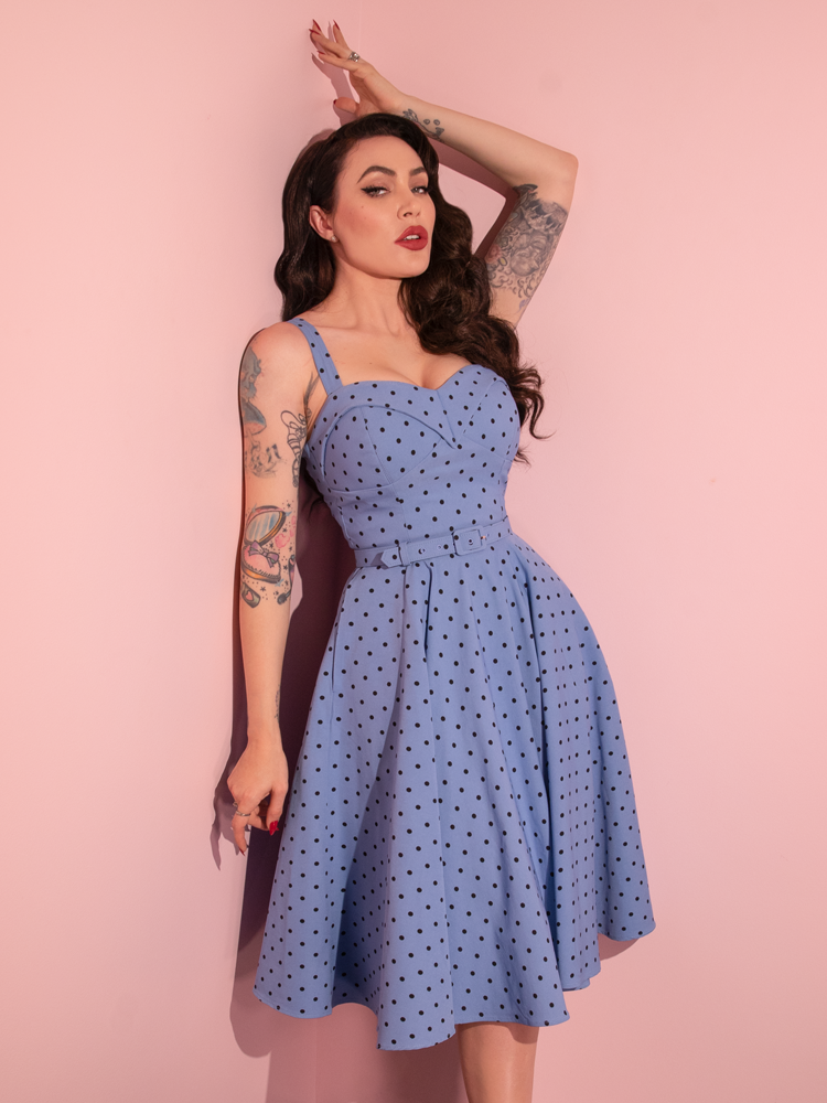 Micheline Pitt leans against the wall while modeling the Maneater Swing Dress in Sunset Blue Polka Dot.
