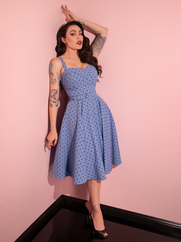 Micheline Pitt leans against the wall while wearing the Maneater Swing Dress in Sunset Blue Polka Dot from retro style clothing brand Vixen Clothing.