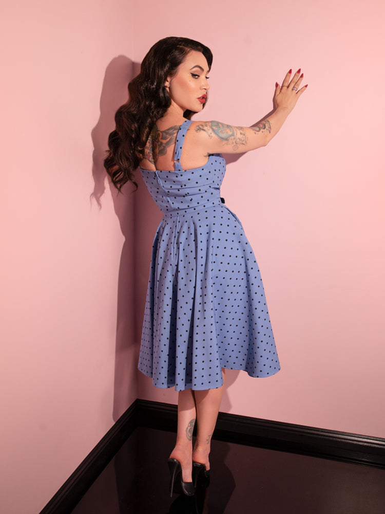 Micheline Pitt faces away from the camera, but looks back over her shoulder, while modeling the Maneater Swing Dress in Sunset Blue Polka Dot.