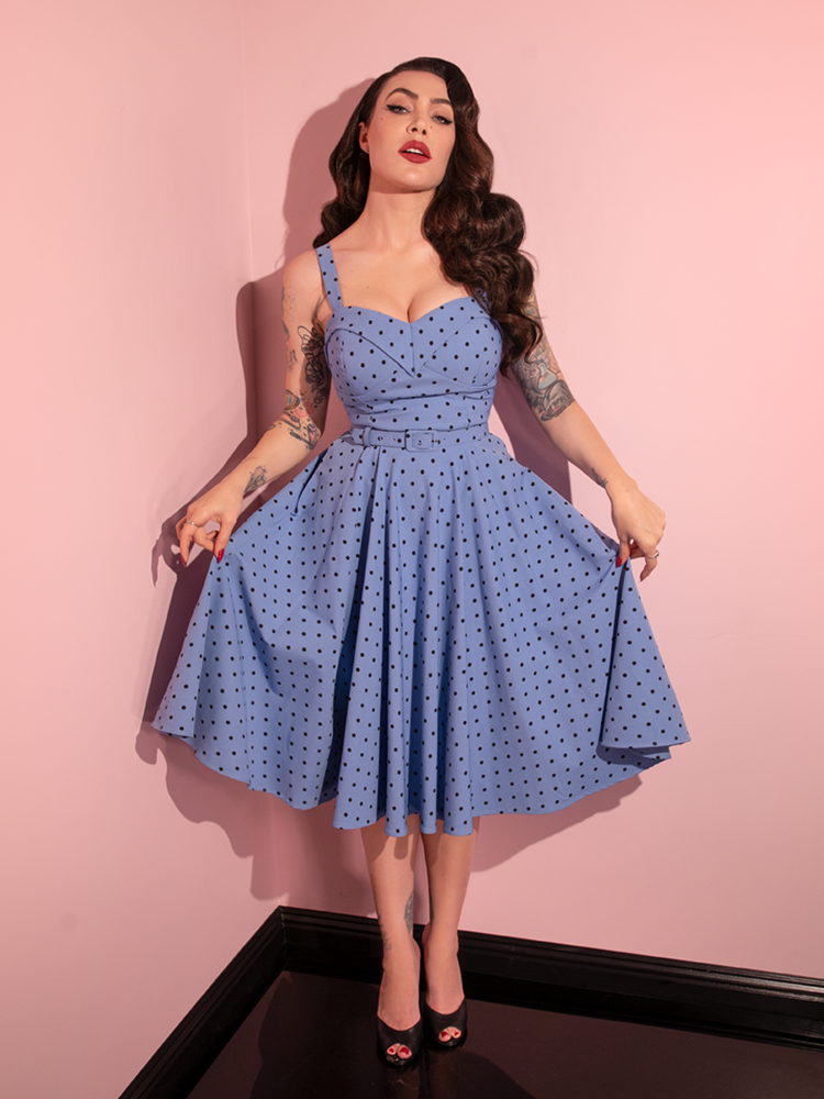 Full length shot of Micheline Pitt holding out the skirt section of the Maneater Swing Dress in Sunset Blue Polka Dot to show off the cute print.