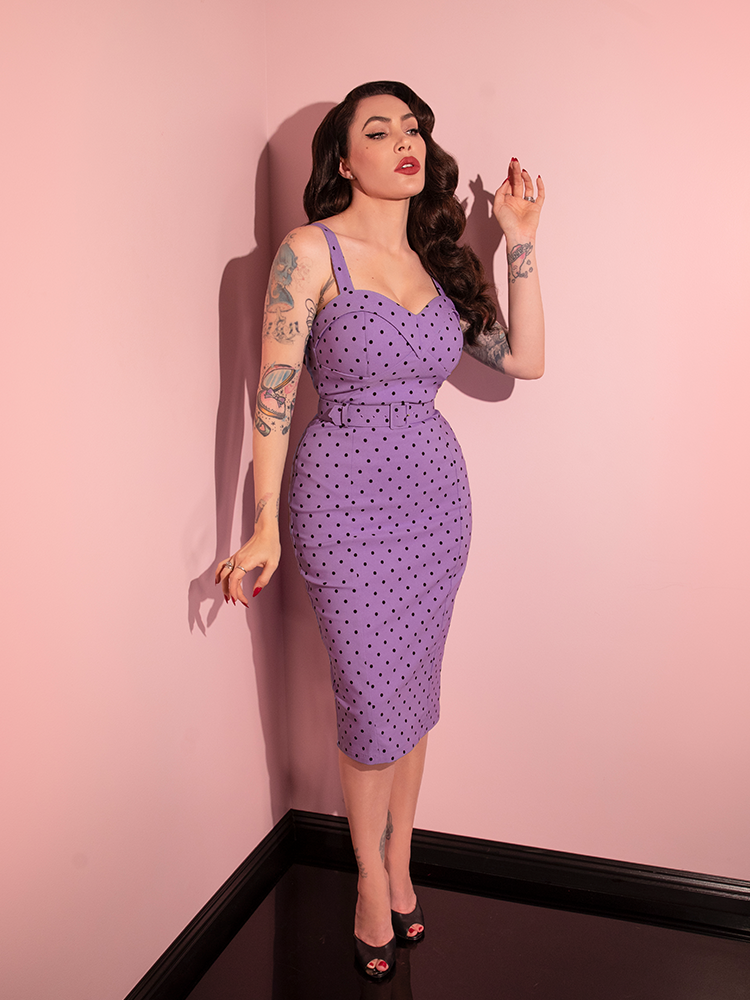 Standing in the corner of a pink showroom wearing the Maneater Wiggle Dress in Sunset Purple Polka Dot, Micheline Pitt poses with her arms outstretched.