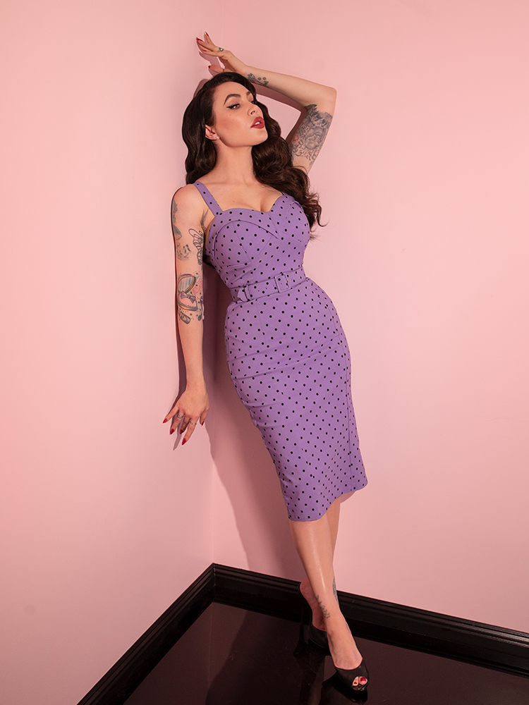 Micheline Pitt leaning against the wall while wearing the Maneater Wiggle Dress in Sunset Purple Polka Dot with open-toed heeled shoes.