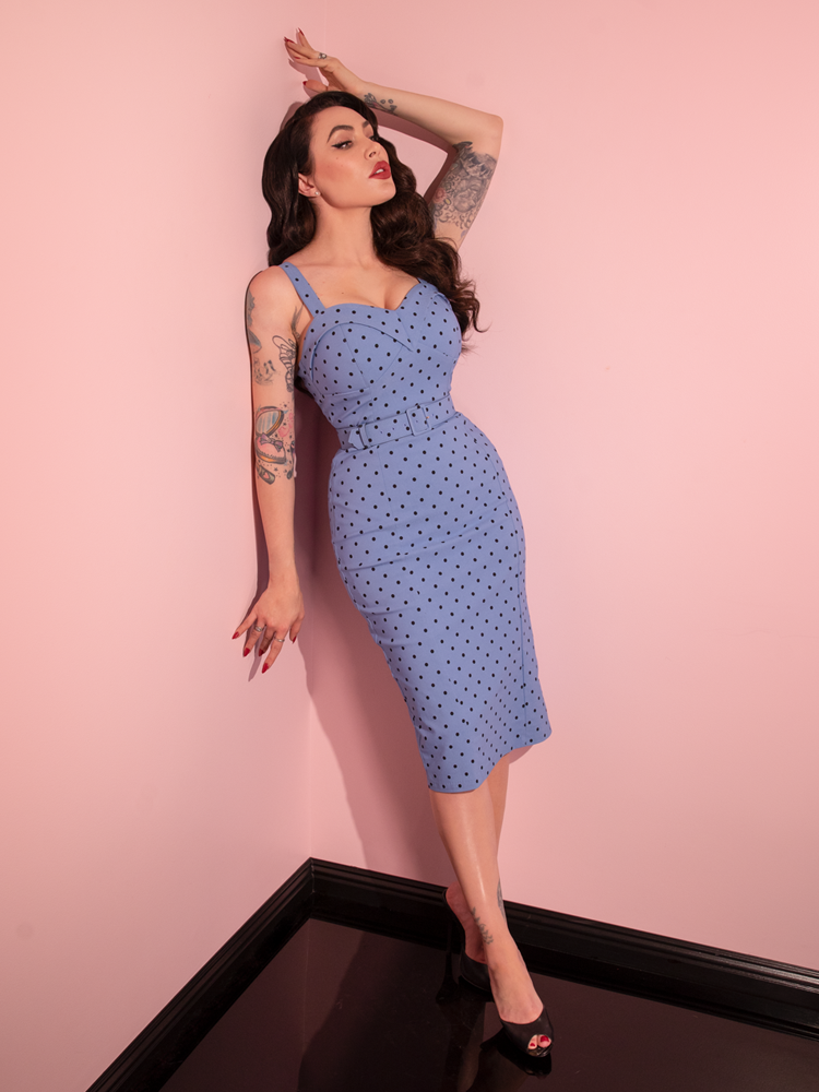 Micheline Pitt leans against the wall and looks away from the camera while wearing the Maneater Wiggle Dress in Sunset Blue Polka Dot.