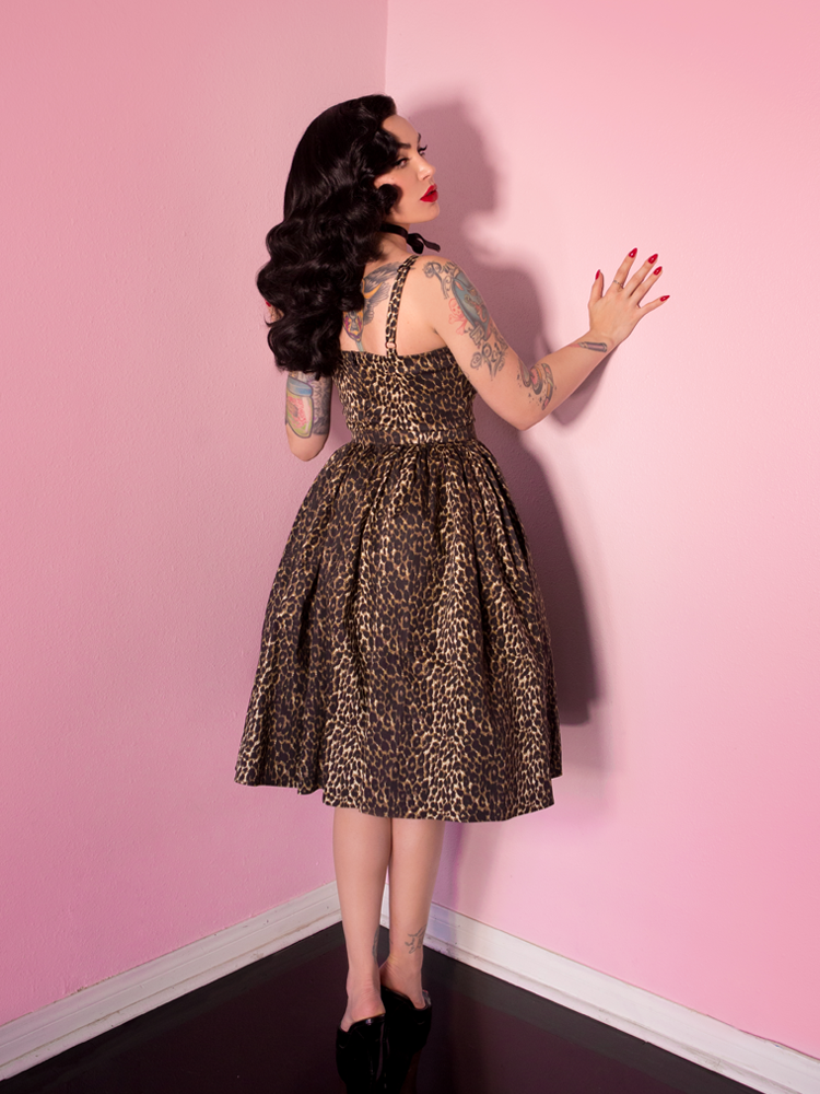 Micheline Pitt leaning against wall and looking over her shoulder, wearing a sweetheart swing dress in a retro style wild leopard print.