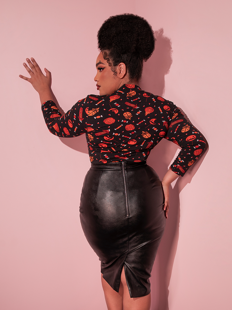 Female model facing away from the camera to show off the back of the TRICK R TREAT™ Bad Girl 3/4 Sleeve Top in Candy Corn Novelty Print tucked into her black vegan leather skirt.