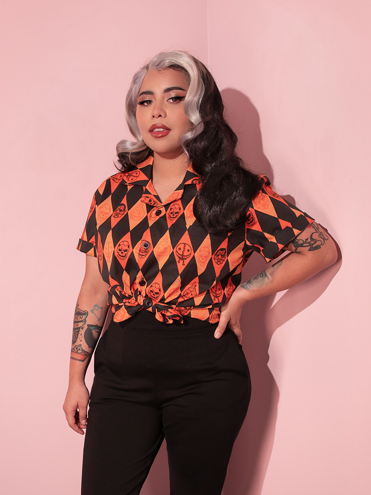 The TRICK R TREAT™ Button Up Shirt in Halloween Harlequin Print being worn by a female model with silver and brown hair and black cigarette pants.