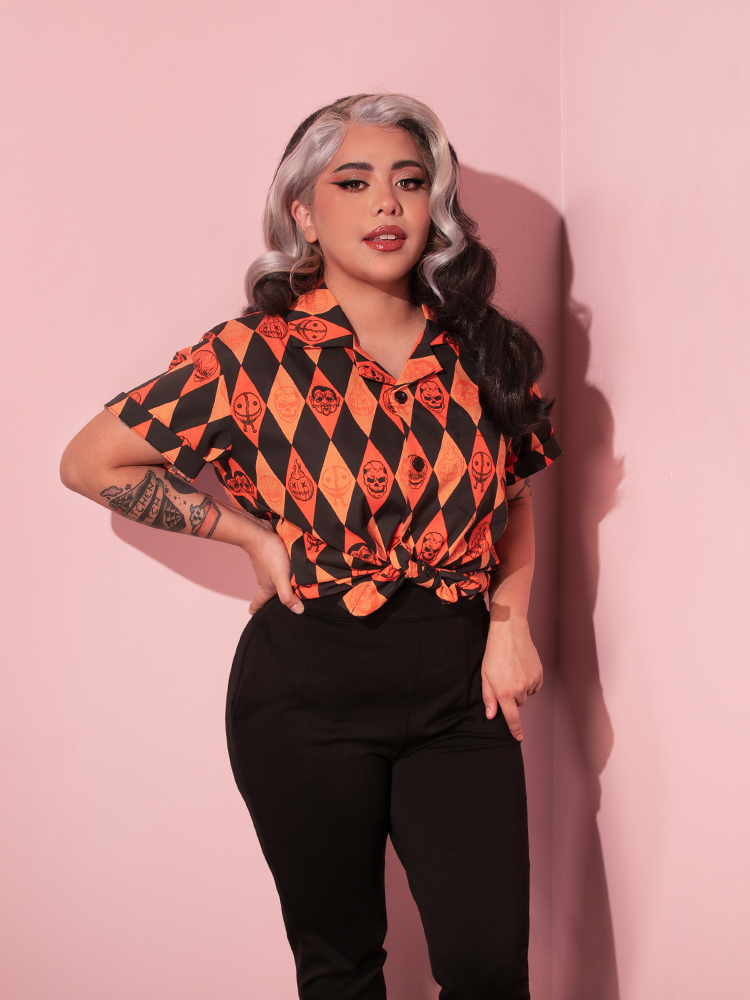 Model posing with one hand resting on her hip while wearing the TRICK R TREAT™ Button Up Shirt in Halloween Harlequin Print paired with a pair of black cigarette pants.