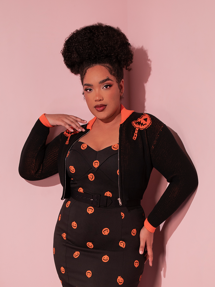 Model Ashleeta wearing a spooky Halloween inspired outfit including the TRICK R TREAT™ Flaming Pumpkin Cropped Knit Jacket over a retro style black dress with pumpkin print.