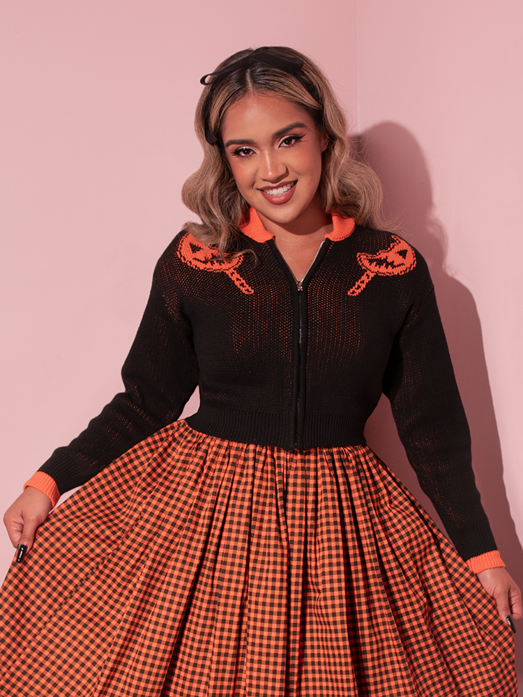 Female Vixen model wearing the TRICK R TREAT™ Flaming Pumpkin Cropped Knit Jacket with a orange and black gingham skirt.