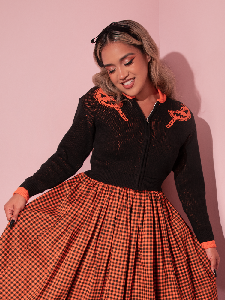 Female model smiling and pulling out the sides of her skirt to show off the gingham print along with the TRICK R TREAT™ Flaming Pumpkin Cropped Knit Jacket she's modeling.
