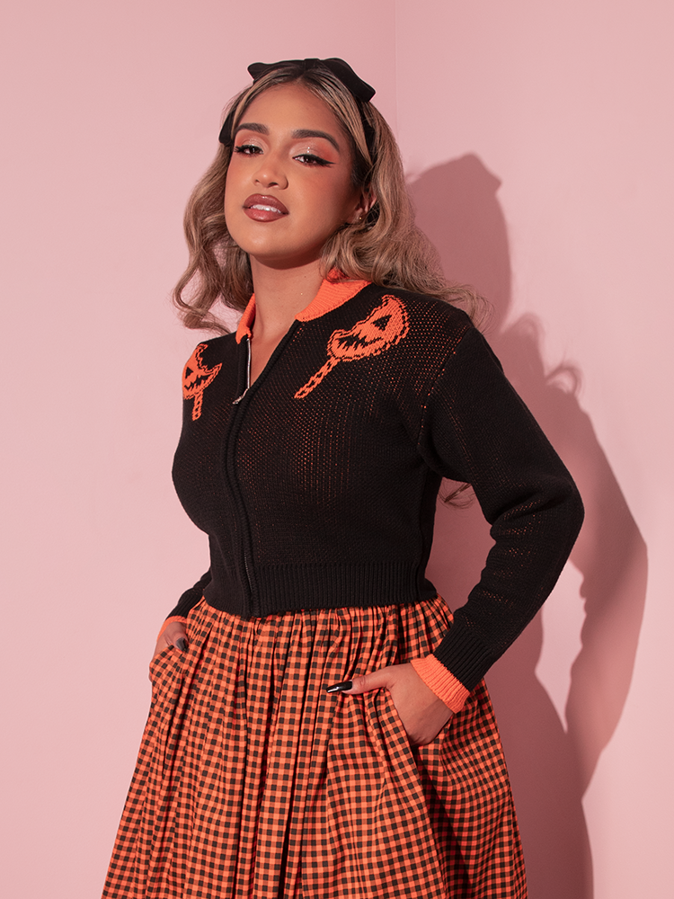 Model tucking her hands into the pockets of her gingham skirt while also wearing the TRICK R TREAT™ Flaming Pumpkin Cropped Knit Jacket to make the perfectly spooky retro style outfit.