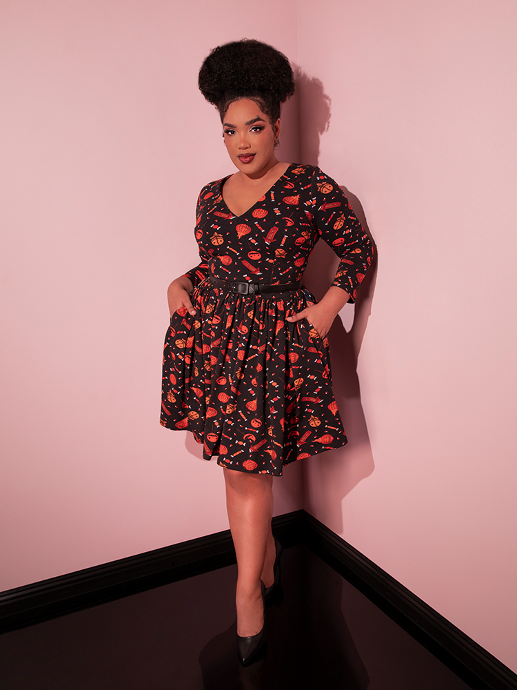 Model tucking her hands into the side pockets of the TRICK R TREAT™ Deadly Swing Dress in Candy Corn Novelty Print from retro clothing company Vixen Clothing.