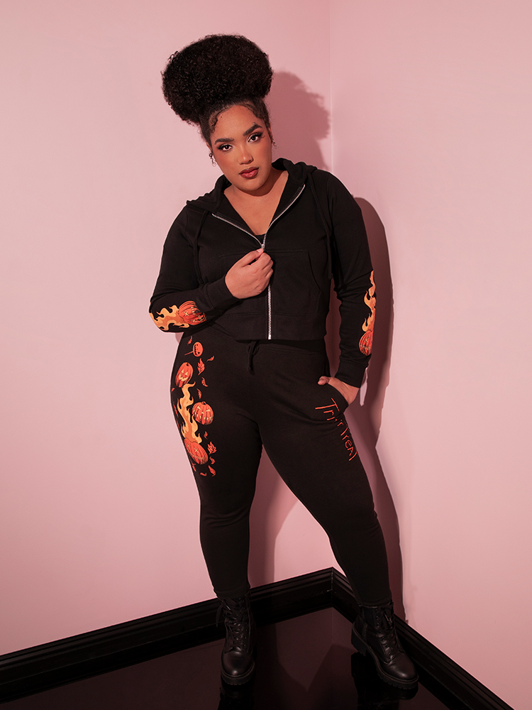 Model standing with on hand tucked into the pocket of her TRICK R TREAT™ Jack O' Lantern Sweatpants while zipping up her black matching hoodie - all items from Vixen Clothing.