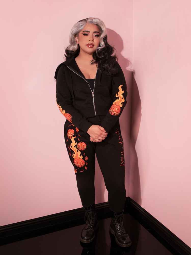 Model standing with her hands clasped in front of her waist while wearing an all black outfit including the TRICK R TREAT™ Jack O' Lantern Sweatpants with matching black hoodie.