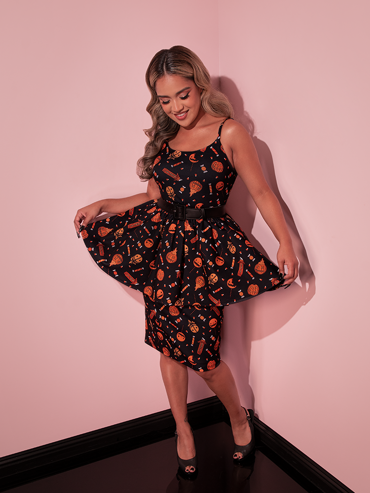 Model hold out the sides of the TRICK R TREAT™ Peplum Wiggle Dress in Candy Corn Novelty Print while looking down at the spooky and festive print.