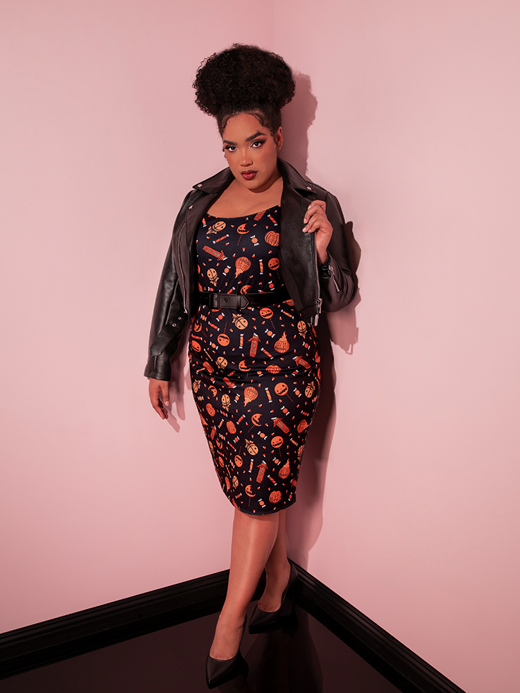 Female model wearing the TRICK R TREAT™ Peplum Wiggle Dress in Candy Corn Novelty Print with a leather jacket and matching vegan leather belt.
