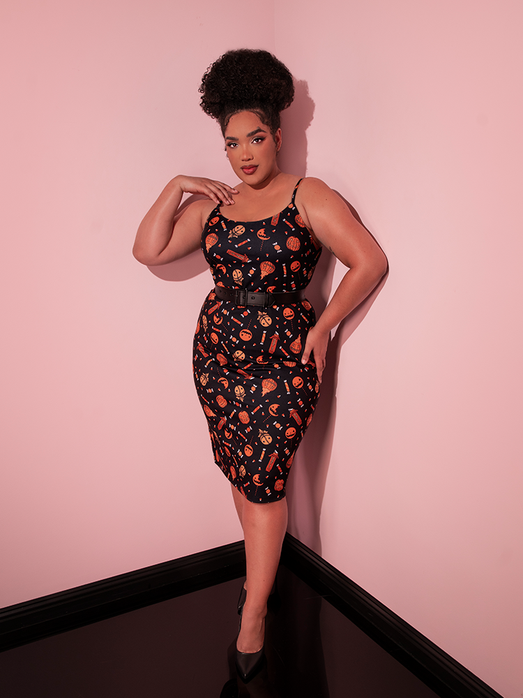 Model posing in the corner of her pink showroom wearing the TRICK R TREAT™ Peplum Wiggle Dress in Candy Corn Novelty Print from retro dress maker Vixen Clothing.