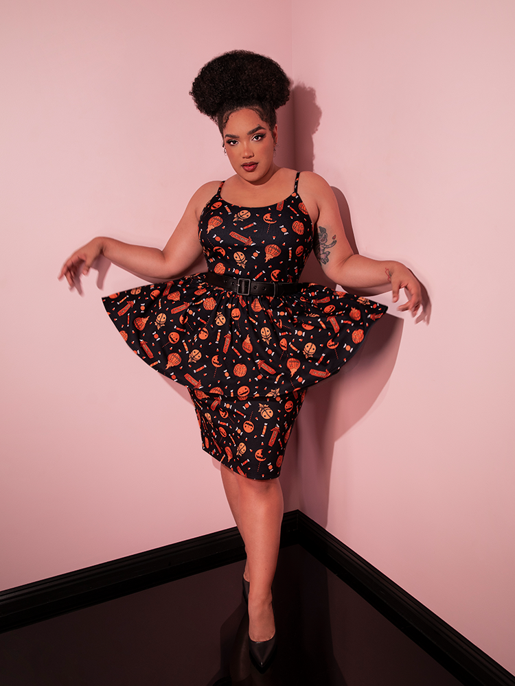 Model pulls out the sides on the skirt section of the TRICK R TREAT™ Peplum Wiggle Dress in Candy Corn Novelty Print to show off the spooky and cute Halloween print.