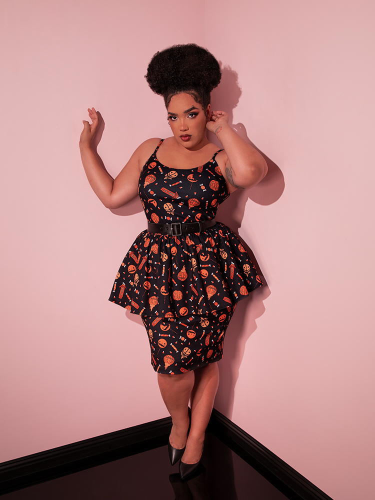 Model posing in the corner of a pink showroom while wearing the TRICK R TREAT™ Peplum Wiggle Dress in Candy Corn Novelty Print with black heeled shoes.