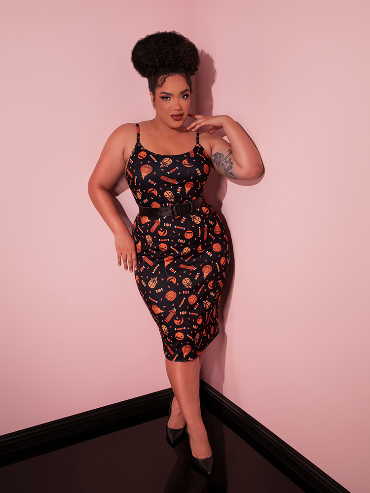 Model leaning back against a wall while wearing the TRICK R TREAT™ Peplum Wiggle Dress in Candy Corn Novelty Print with matching black shoes.