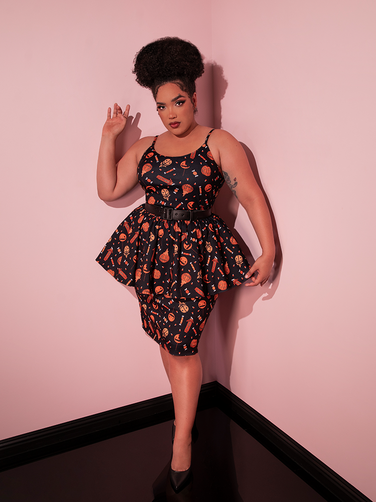 Model posing in the corner of a room with one hand pulling out the skirt section of the TRICK R TREAT™ Peplum Wiggle Dress in Candy Corn Novelty Print to show off the spookily festive print.
