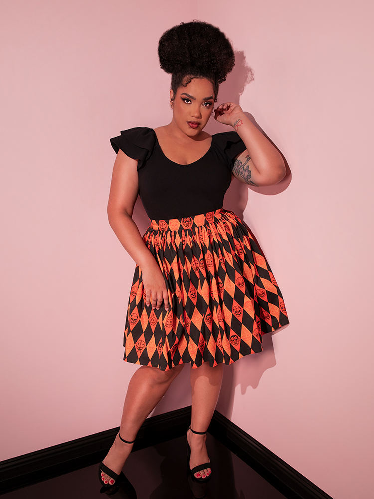 Model posing with one hand next to her head and the other resting on her leg, stands while wearing the TRICK R TREAT™ Skater Skirt in Halloween Harlequin Print with a paired black top.