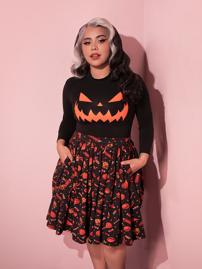 Female model poses in the corner of a pink room while wearing the TRICK R TREAT™ Skater Skirt in Candy Corn Novelty Print with her hands tucked into the side pockets.